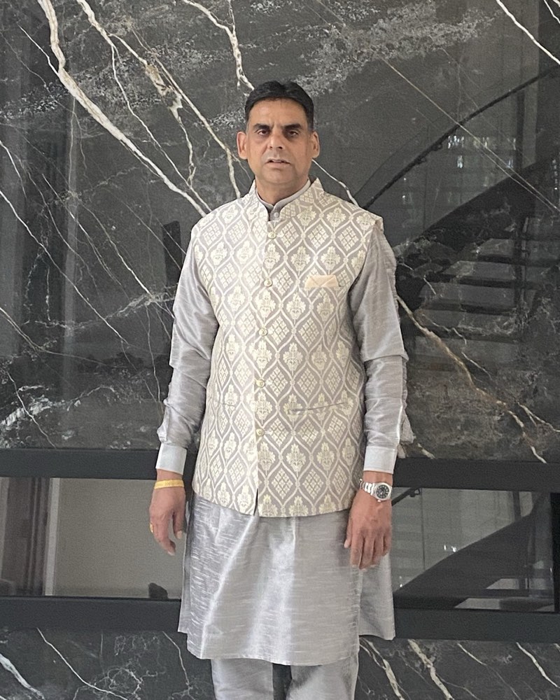 Sanjeev Grover in a silver kurta and brocade vest, standing in front of a marble wall